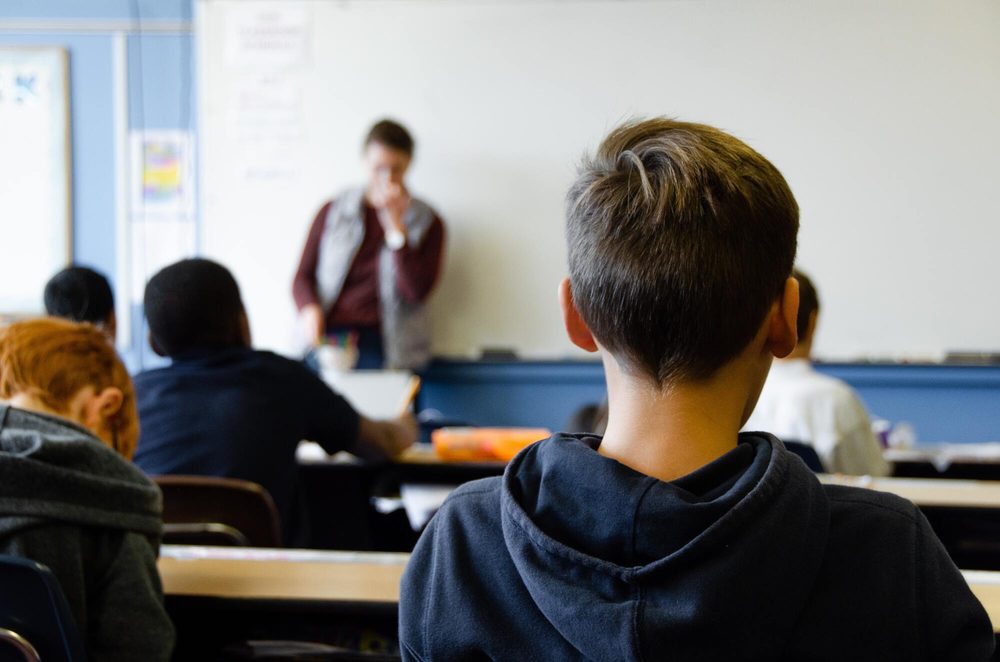 Un alumno de espaldas en clase, con el resto del aula y la profesora desenfocados al fondo